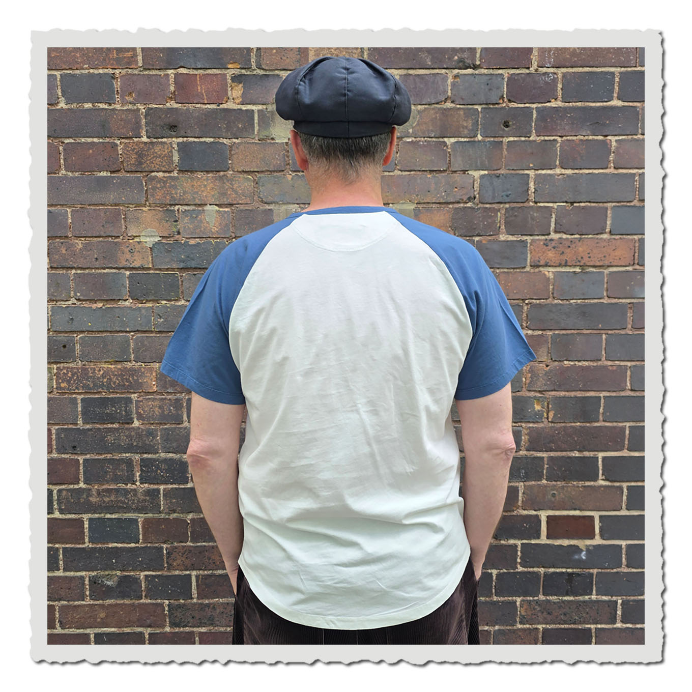 Rückansicht einer Person im weißen T-Shirt mit blauen Ärmeln vor Backsteinwand im Freien.